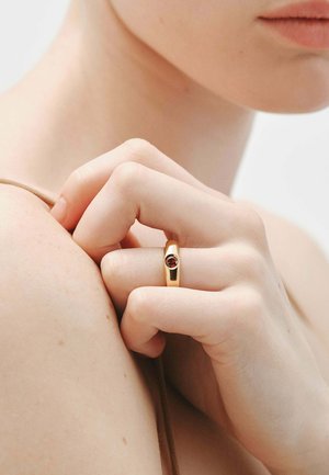 Anillo de oro con un acabado liso y pulido, que presenta una piedra preciosa roja en el centro. La mano reposa sobre un fondo de color neutro.