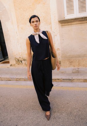 Kobieta idąca ulicą w czarnym topie bez rękawów, szerokich czarnych spodniach, lekkim szaliku na szyi, brązowych spiczastych butach, niosąca plecioną torbę na ramię.