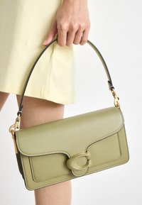 Ręka trzyma jasnozieloną skórzaną torebkę z złotym zapięciem i czarnym paskiem na tle jasnopomarańczowej spódnicy.