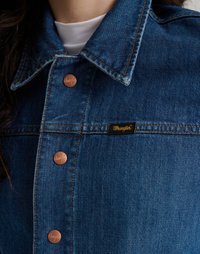 Τζάκετ τζιν σε κλασικό μπλε χρώμα, με κουμπί μπροστά και χάλκινα κουμπιά, καθώς και λογότυπο Wrangler στο αριστερό στήθος.