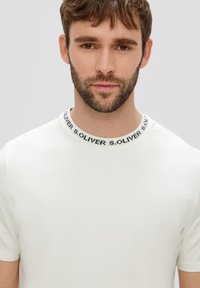 Biała koszulka z okrągłym dekoltem, z czarnym detalem z bandą "S.OLIVER" wokół kołnierza. Krótkie rękawy, miękka tekstura.