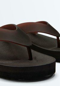 Close-up van zwarte en donkerbruine teenslippers met brede teenbandjes en gestructureerde voetbedden op een witte achtergrond.
