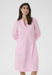 Rožinė medvilninė suknelė su apykakle, ilgomis pūstomis rankovėmis ir laisvu siluetu, besitęsiančiu virš kelių. Paprastas mygtukų akcentas.
