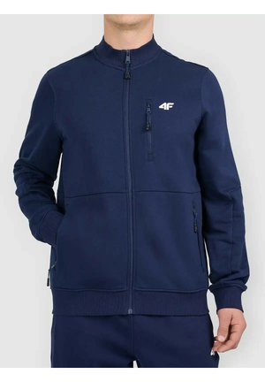 Mężczyzna w sportowej kurtce granatowej zapinanej na zamek z logo 4F, kieszenią na piersi na zamek i bocznymi kieszeniami, stojący na białym tle.