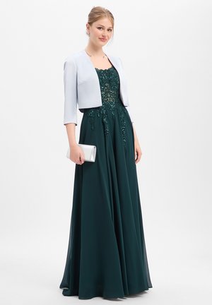 Junge Frau trägt ein dunkelgrünes verziertes Kleid und eine hellgraue kurze Jacke, hält eine silberne Clutch und steht vor weißem Hintergrund.
