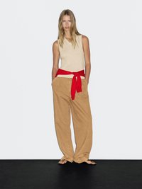 Donna con lunghi capelli biondi che indossa un top beige senza maniche, pantaloni beige larghi, un maglione rosso annodato in vita e sandali neri, in piedi contro un muro bianco.