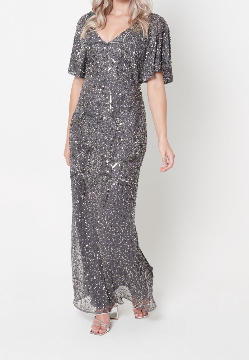 Femme portant une robe longue grise à sequins avec de courtes manches volantes et des sandales argentées à talons hauts sur fond blanc.