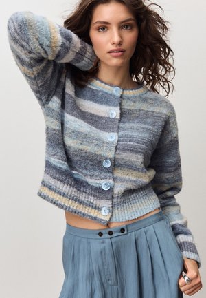 Giovane donna con capelli mossi indossa un cardigan a righe blu-grigio con bottoni e una gonna plissé blu a vita alta, posando con un braccio alzato.