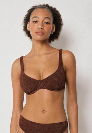 Femme aux cheveux bouclés naturels portant un haut de bikini texturé marron et un bas assorti, sur un fond clair uni.