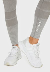 Graue Leggings mit einem gerippten Muster und "SQUATPROOF"-Branding. Weiße Sportschuhe besitzen ein Mesh-Obermaterial und ein Design mit drei Streifen.