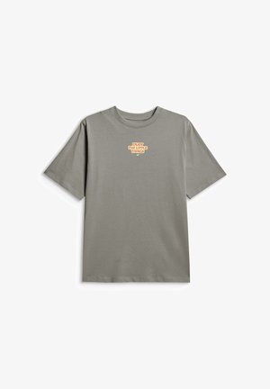 Młoda osoba z długimi blond włosami, ubrana w szarą oversize koszulkę z napisem "Enjoy The Little Things", czarne spodnie i kolorowe sneakersy.
