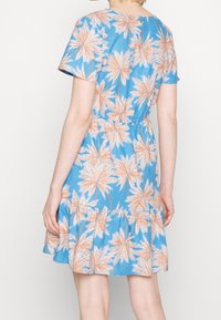 Femme portant une robe bleue à manches courtes avec des motifs de palmiers beiges, vue de dos sur fond uni.