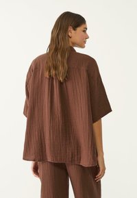 Donna dai lunghi capelli castani che indossa un top corto a maniche larghe, marrone e strutturato, insieme a pantaloni abbinati, vista da dietro su uno sfondo bianco.