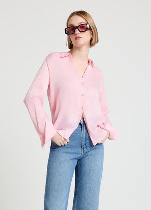 Femme portant une chemise rose clair boutonnée, un jean bleu et de grandes lunettes de soleil carrées foncées, debout devant un fond clair uni.