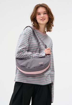 Junge Frau trägt ein pink-grünes gestreiftes Langarmshirt und eine graue und pinke Umhängetasche mit verstellbarem Schultergurt.