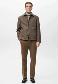 Braune Wolljacke mit Kragen, zwei Vordertaschen und Knopfverschluss, kombiniert mit braunen Hosen und schwarzen Schuhen, stehend vor einem schlichten Hintergrund.