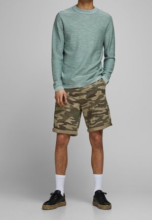 Hellgrünes Langarmshirt mit strukturiertem Muster, kombiniert mit braunen Camouflage-Shorts. Schwarze Sneakers, weiße Socken. Lässiges Outfit-Design.
