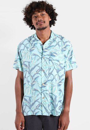 Homme aux cheveux bouclés portant une chemise à manches courtes bleu clair à boutons avec un motif de feuilles et un pantalon noir.