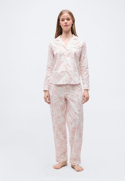 Νεαρή γυναίκα standing barefoot wearing pale pink floral long-sleeve button-up pajama top and matching loose pants σε λευκό φόντο.