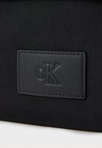 Črnen usnjen nahrbtnik z teksturiranim usnjenim oznako z odtiskanim logotipom "cK", dopolnjenim z detajli šivanja okoli oznake.