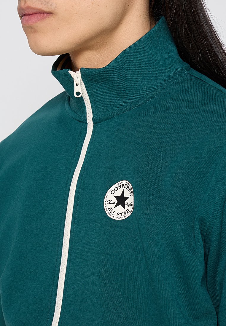 Giacca a zip color teal con colletto alto, caratterizzata da una zip bianca e una patch rotonda in bianco e nero sul petto. Tessuto morbido.