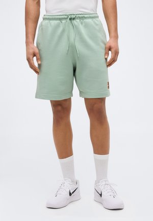 Férfi világoszöld, húzózsinóros rövidnadrágban, fehér bokazokniban és fehér Nike tornacipőben, egyszínű háttér előtt állva.