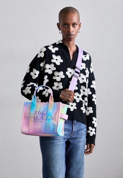 Väike, mitmevärviline kott siledast materjalist, millel on tekst "THE TOTE BAG" ja lilla rihm. Kandmisel must lilleline sviiter.