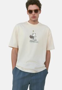 Camiseta de manga corta color crema con un gráfico de Snoopy sosteniendo una raqueta de tenis. Textura suave, corte relajado, combinada con pantalones azules con estampado.