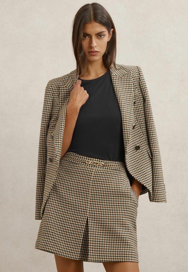 Rudas ir kreminis languotas švarkas, derinamas su juodu viršumi ir to paties rašto languota sijonu. Turi dvigubą sagų eilę ir klostytą dizainą.