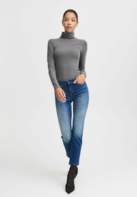 Maglione a collo alto grigio a maniche lunghe abbinato a jeans in denim blu, caratterizzato da un design aderente e una leggera sbiaditura, completato da tacchi neri a punta.