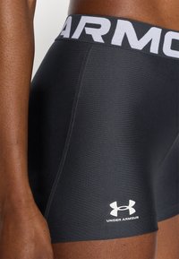 Shorts de sport noirs de Under Armour, avec un logo blanc et une large ceinture. Le tissu a une texture lisse et une coupe ajustée.