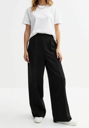 Personne portant un t-shirt blanc, un pantalon noir à jambes larges, des baskets blanches et un collier chaîne en or, debout devant un fond uni.