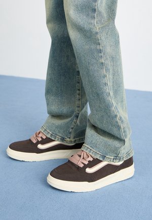 Bruine en zwarte sneakers met roze veters en een witte zool, gecombineerd met lichtblauwe spijkerbroeken. Sneakers hebben een gebogen zijstreep.