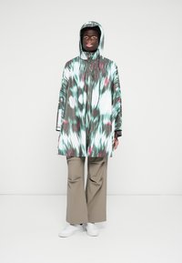 Multicolored waterproof poncho with a hood, featuring a tie-dye pattern in shades of green, white, and pink, paired with loose khaki pants.