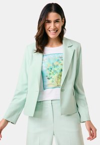 Mintgrüner, taillierter Blazer mit strukturiertem Finish, ein Knopfverschluss und Reverskragen, kombiniert mit einem weißen T-Shirt mit Grafik darunter.