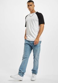 Vit och svart kortärmad raglan-t-shirt i kombination med ljusblå jeans och vita sneakers. Modellen står vänd något åt sidan.