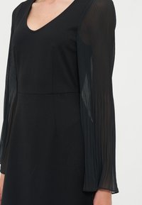 Robe noire avec encolure en V et manches longues transparentes présentant des plis verticaux sur une personne à la peau claire.