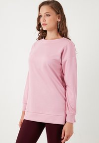 Felpa rosa a maniche lunghe con scollo a girocollo, texture morbida e vestibilità rilassata, con un orlo leggermente più lungo sul retro.