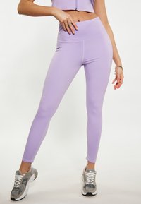 Leggings a vita alta color lilla realizzati in tessuto elastico, caratterizzati da una superficie liscia e dettagli con cuciture, abbinati a sneakers grigie.