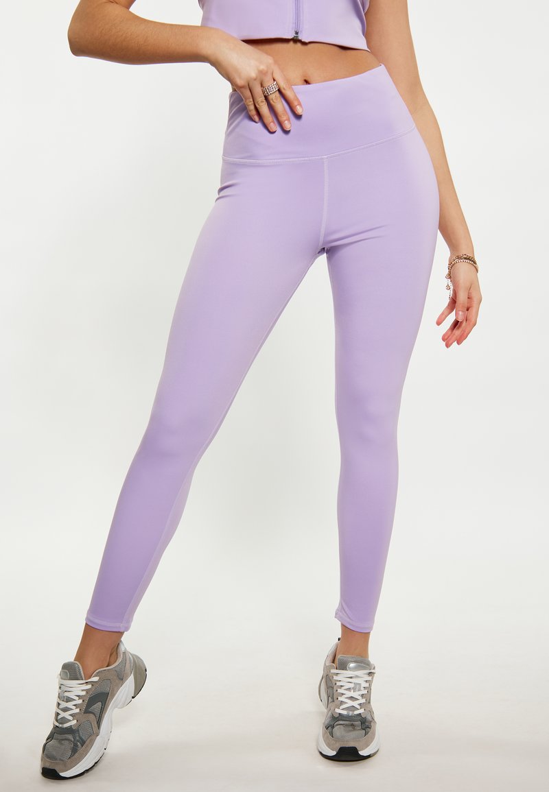 Leggings a vita alta color lilla realizzati in tessuto elastico, caratterizzati da una superficie liscia e dettagli con cuciture, abbinati a sneakers grigie.