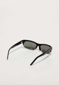 Lunettes de soleil noires rectangulaires avec verres foncés, monture en plastique glossy, branches fines et accents dorés sur les tempes.