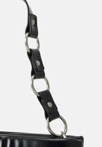 Anse en cuir noir avec des anneaux en métal argenté reliés par des sections en cuir, fixée à la fermeture éclair d'un sac noir texturé.