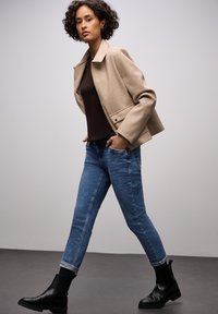 Giacca di lana beige con colletto strutturato, indossata sopra una maglia marrone scuro, abbinata a jeans di denim blu e stivaletti neri.
