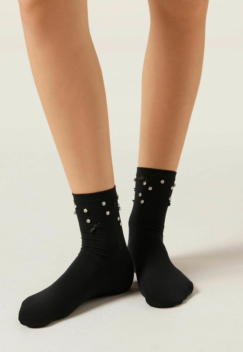 Czarne skarpety wykonane z gładkiego materiału, zdobione elementami dekoracyjnymi, takimi jak małe perły i kokardy na kostce. Klasyczny, dopasowany kształt.