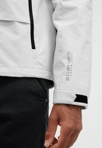 Chaqueta blanca con un acabado texturizado, con cremallera negra y acentos negros. Logotipo impreso en la manga. Lleva puesta sobre pantalones negros.