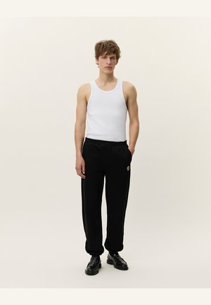 Top sin mangas blanco con pantalones deportivos negros que presentan un parche de logo, corte recto, cinturilla elástica y zapatos negros. Un atuendo simple y casual.