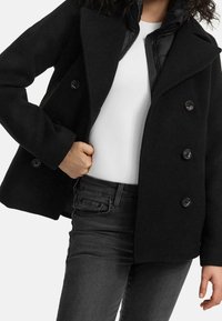 Černý dvouřadý kabát s plášťovým límcem a čtyřmi knoflíky. Nosit na bílém tričku, v kombinaci s tmavě šedými džíny. Hladká textura.