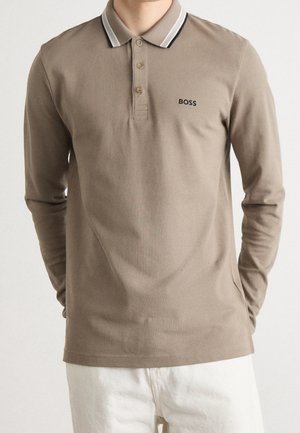 Homme portant un polo beige à manches longues avec un col rayé noir et blanc et le logo "BOSS" sur la poitrine gauche, associé à un pantalon blanc.