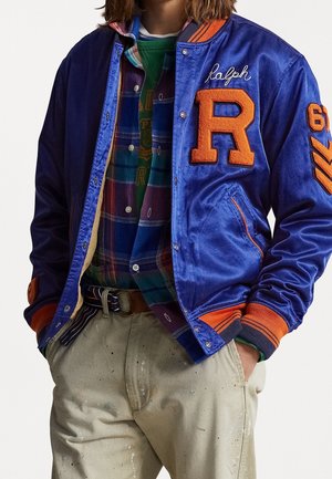 Person trägt eine blaue und orangefarbene Varsity-Jacke mit großem "R" und "Ralph" Stickerei, kariertes Hemd, grünen Pullover und beige, mit Farbflecken gesprenkelte Hose.