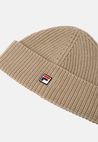 Beige neulottu pipa, jossa on ribbidattu pinta ja käännetty reuna. Edessä pieni monivärinen Fila-logopatch.